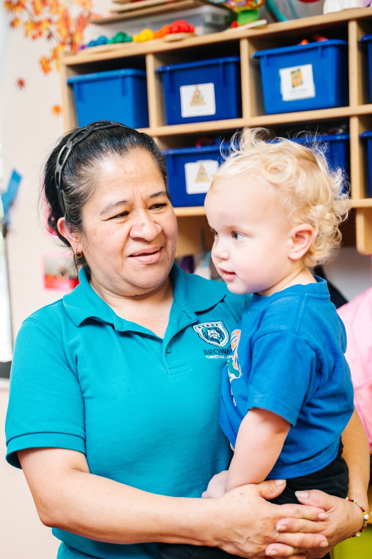 Teacher holding a toddler in a warm, colorful classroom